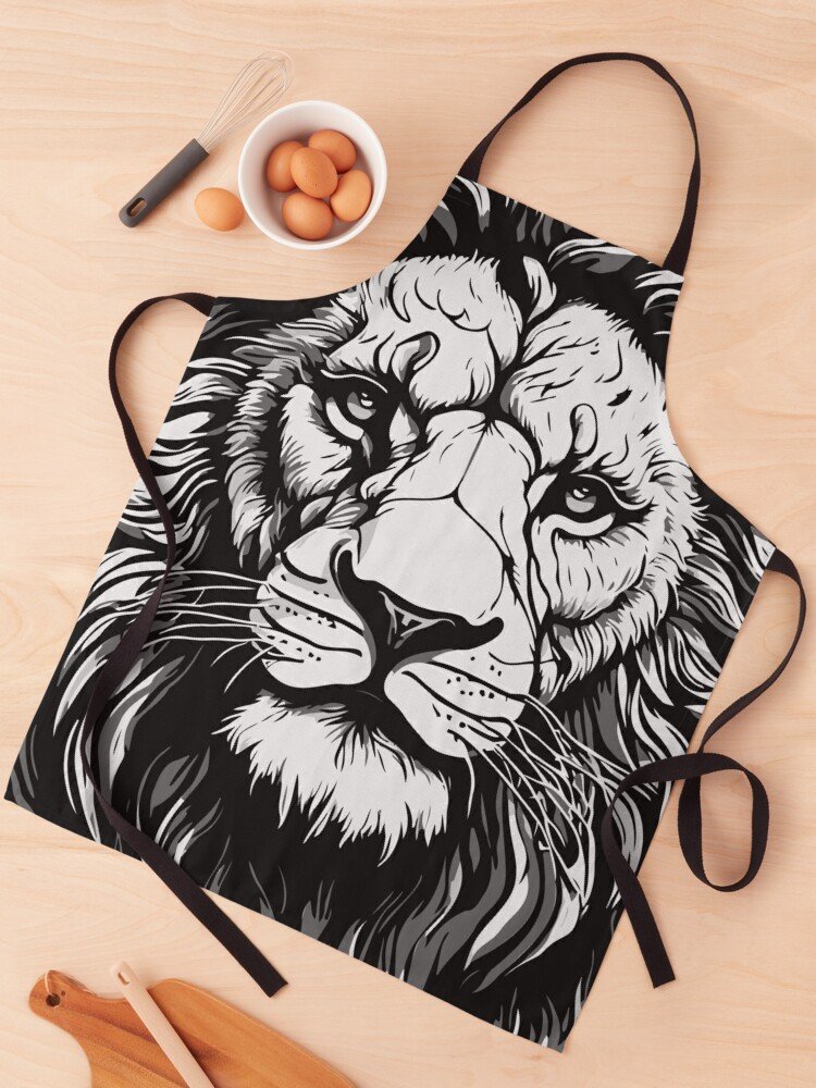 Delantal negro con ilustración de león: Delantal negro con una impresión grande de la cara de un león en blanco y negro, colocado sobre una mesa con utensilios de cocina.